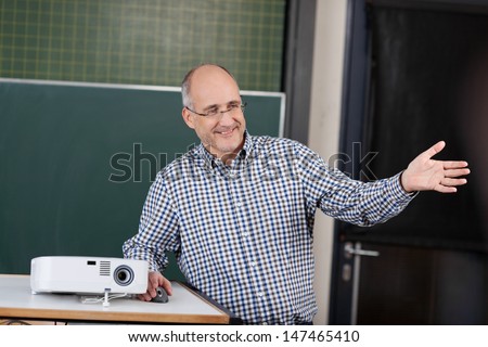 Professor at a university giving a presentation standing in front of the chalkboard with a projector gesturing with his hand for the class to look at something