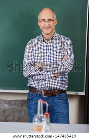 Middle-aged male teacher standing with his arms folded against the blackboard in chemistry class with scientific glassware on the table in front of him