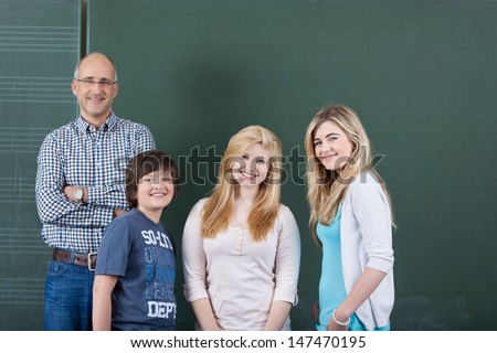 Successful team at school with a smiling confident male teacher posing with two teenage girls and boy from his class in front of the blackboard