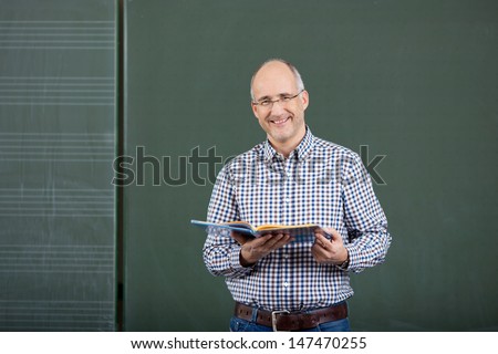 Friendly male teacher teaching form a set of class notes standing in front of a blank green blackboard with copyspace