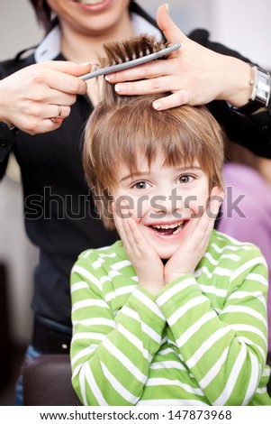 Cute young child at the hairdresser laughing with his hands to his face as the stylist cuts his hair with scissors