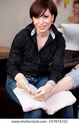 Pedicurist in a nail studio or beauty salon sitting with a clients bare foot on her lap smiling up at the camera
