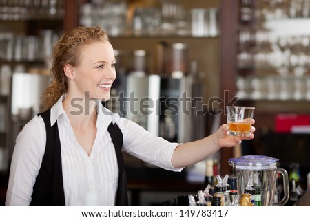 Smiling beautiful young barmaid serving alcohol in a tumbler to a client across the top of the bar counter