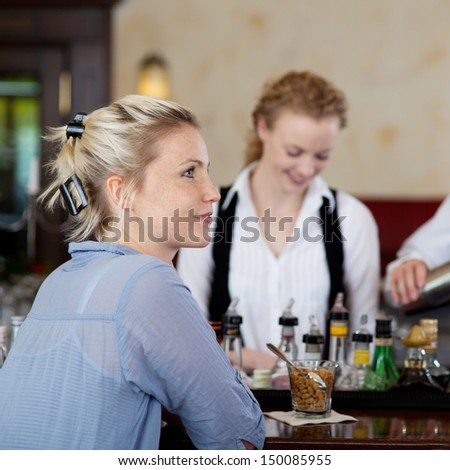 Beautiful young woman sitting at a bar in a nightclub or restaurant with a waitress busy in the background