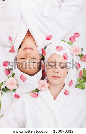 Two women friends relaxing in a spa lying head to head on their backs with their eyes closed and rose petals and fresh pink roses scattered around their faces