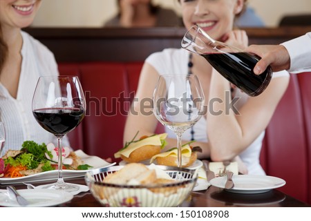 Waiter pouring red wine from a carafe at a restaurant for two young women seated at a table with their food