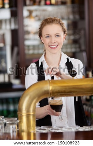 Smiling attractive young barmaid standing behind the bar serving dark draft beer