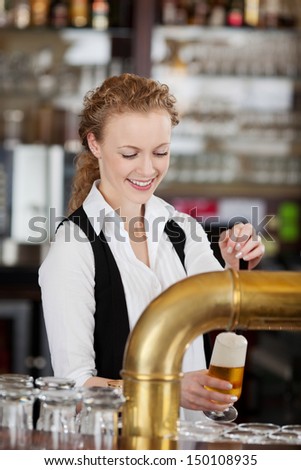 Happy barmaid pouring draft beer standing behind the counter in a bar or pub filling a pint glass