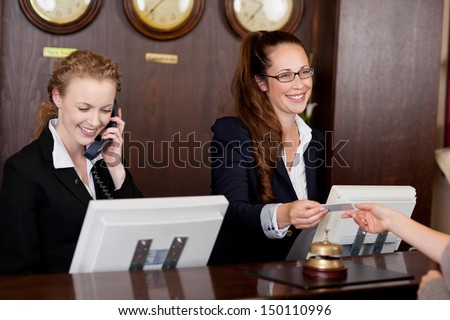 Two beautiful young stylish receptionists at a reception desk, one talking on the telephone and the other handing a card to a customer