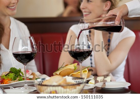 Two women enjoying red wine sitting eating a meal in a restaurant and chatting while the waiter replenishes their glasses