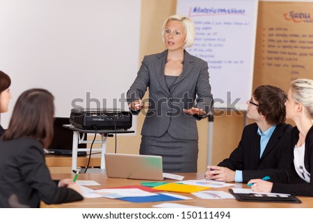 Woman Sung A Projector Giving A Corporate Training Class To A Group Of Young Businesspeople Around A Table Gesturing With Her Hands As She Explains Something