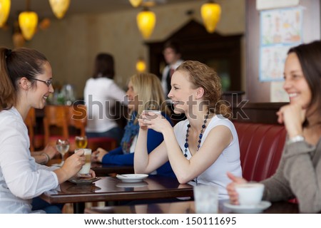 Guests Enjoying Coffee In A Restaurant With Focus To Two Stylish Young Woman Sitting At A Table Together