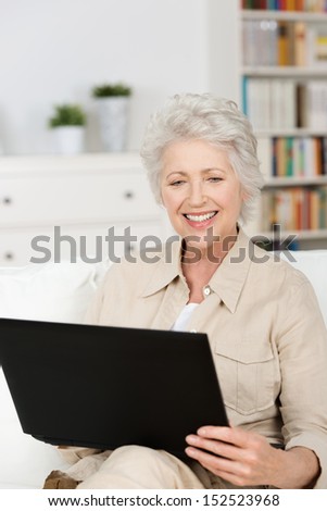 Senior woman using a laptop at home sitting on the sofa in the living room reading information on the screen with a smile