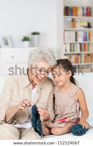Senior Lady Teaching Her Granddaughter To Knit As They Relax Together On The The Sofa Laughing Together With Enjoyment