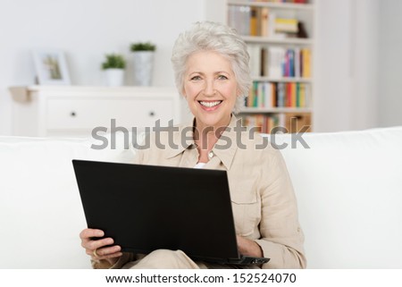 Senior Woman Working An A Laptop Balanced On Her Lap As She Relaxes On A Comfortable Sofa At Home Smiling At The Camera