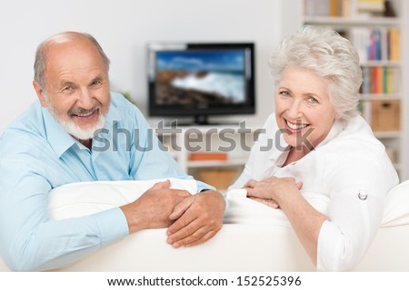 Happy Friendly Elderly Couple Relaxing In Their Living Room In Front Of The Television Turning To Smile At The Camera Over The Back Of The Sofa
