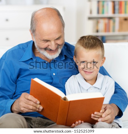 Grandfather reading to his small grandson who is looking up to smile at the camera as they sit arm in arm close together on a sofa in the living room