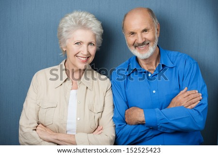 Confident elderly couple with folded arms standing side by side smiling at the camera against a green background