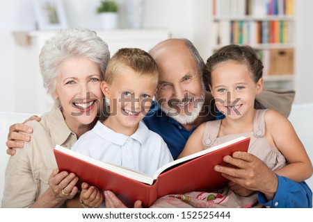 Elderly Couple Reading To Their Grandchildren As They All Sit Close Together On A Sofa