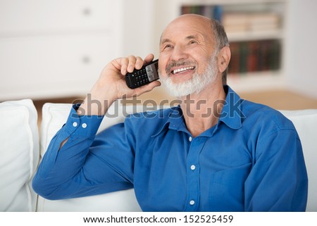 Delighted senior man chatting on a mobile phone while relaxing on a sofa in his living room, natural close up portrait
