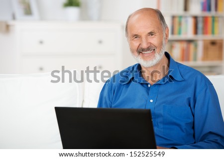 Confident elderly man giving the camera a beautiful smile using a laptop while relaxing on a couch at home in his living room