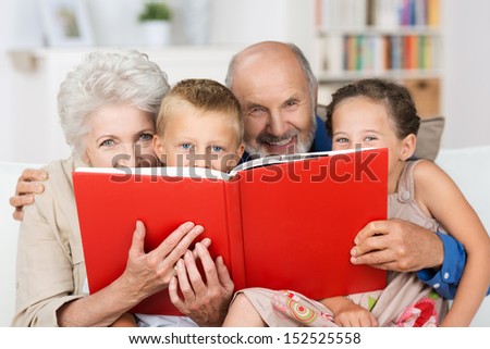 Cute little boy and girl with merry smiling eyes reading with their grandparents peering over the top of the book at the camera