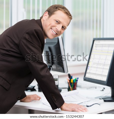 Confident Successful Young Businessman Leaning His Arms On His Desk To Look At Information On His Computer Turning To Smile At The Camera
