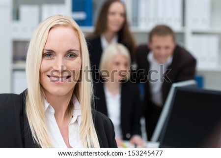Successful confident beautiful businesswoman with a warm smile and long blond hair posing in an office with her colleagues in the background