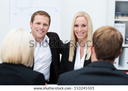 Smiling successful coworkers, a man and woman, in a business meeting sitting side by side facing the camera, view between two people with their backs to the camera