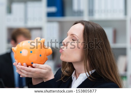 Beautiful young businesswoman standing in her office kissing her piggy bank which she is holding cupped in her hands