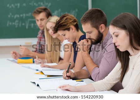 Group Of Diverse Young Students At Work In The Classroom Sitting At A Long Table Working On Their Notes And Studies