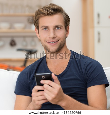 Handsome unshaven casual young man holding a smartphone in his hands and looking at the camera with a smile, indoors portrait