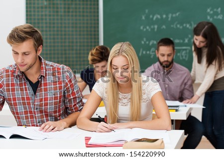 Attractive young man and woman studying together at their desks in a college or university classroom with other students and a blackboard behind them