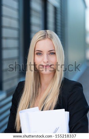 Stylish young female business executive with long blond hair holding a sheaf of paperwork standing outside the office building looking at the camera