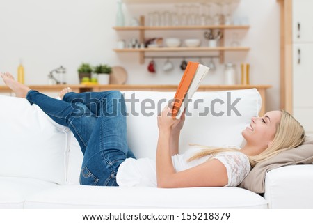 Pretty Young Woman Enjoying Reading A Book At Home Lying On The Sofa Smiling In Pleasure In Casual Clothing