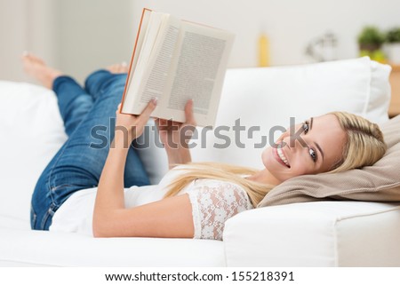 Beautiful woman reading on a sofa relaxing with her bare feet over the arm of furniture turning to give the camera a beaming smile