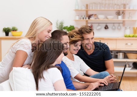 Group of young people sharing a laptop sitting together on a sofa in the living room smiling as one man surfs the internet