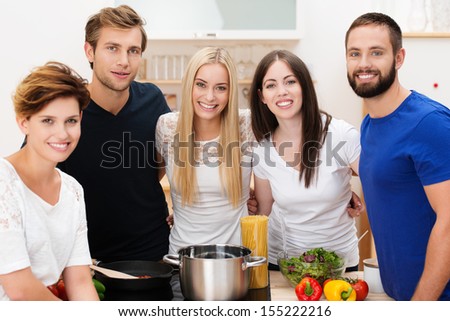 Group of happy young friends preparing lunch standing smiling at the camera grouped around the stainless steel pot and fresh healthy ingredients
