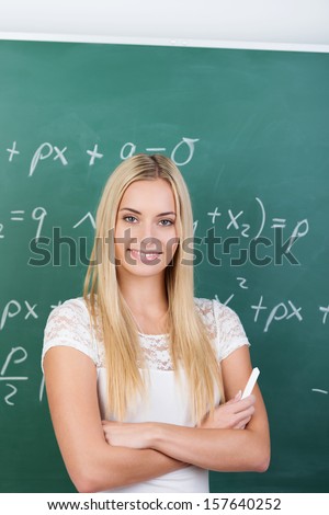 Confident beautiful young female student standing with folded arms in front of a blackboard covered in equations