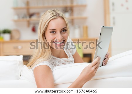 Beautiful vivacious young woman with a tablet computer in her hands leaning over the back of a sofa smiling at the camera