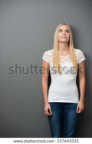 Thoughtful slender young blond woman standing with her arms at her side staring upwards into space against a blank green chalkboard