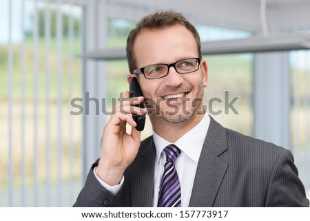 Cheerful businessman wearing glasses having a phone conversation standing in his office listening carefully and smiling