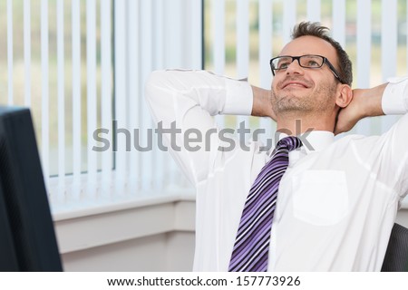 Businessman relaxing at his desk leaning back in his chair with his hands behind his head and a contented smile