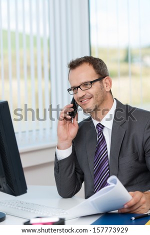 Smiling businessman talking on the phone as he sits at his desk in the office with paperwork in his hand