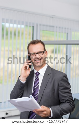 Businessman chatting on his mobile phone in the office standing looking up with a smile while holding paperwork