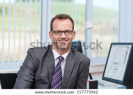 Friendly Businessman Wearing Glasses Sitting At His Desk In The Office Looking At The Camera With A Smile
