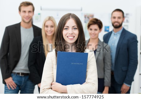 Attractive smiling young businesswoman holding her curriculum vitae in a blue folder as she stands waiting for a job interview with other diverse applicants in the background