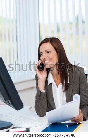 Businesswoman working with paperwork and a computer at her desk smiling and chatting on her mobile phone