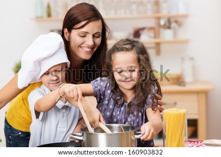 Small Brother And Sister Cooking A Meal Both Stirring The Contents Of The Same Pot Watched Over By Their Laughing Young Mother