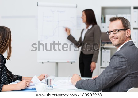 Smiling businessman in a meeting or lecture at the office turning to look at the camera while a businesswoman gives a presentation in the background